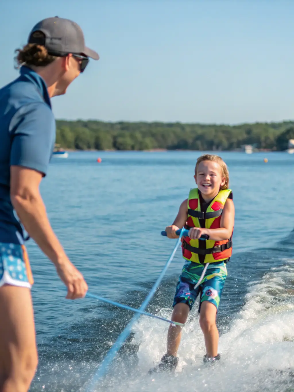 A young child in a life vest smiles brightly while learning to steer a small sailboat with the help of an instructor on a sunny day at ECOLE VOILE DE LION SUR MER.